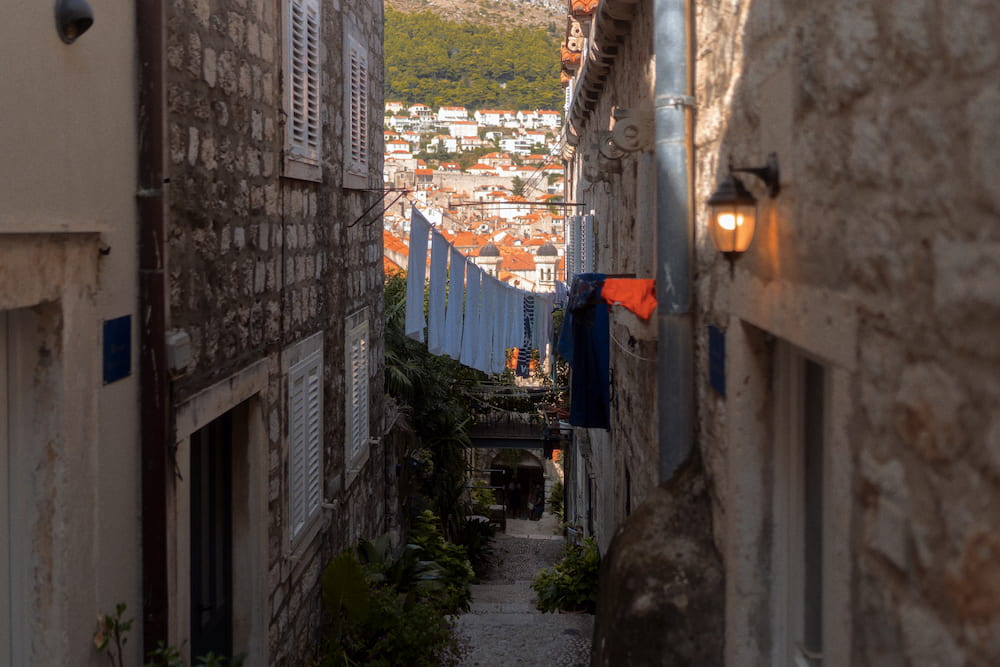 A narrow lane with laundry and a lamp, roofs and a hill at the end.