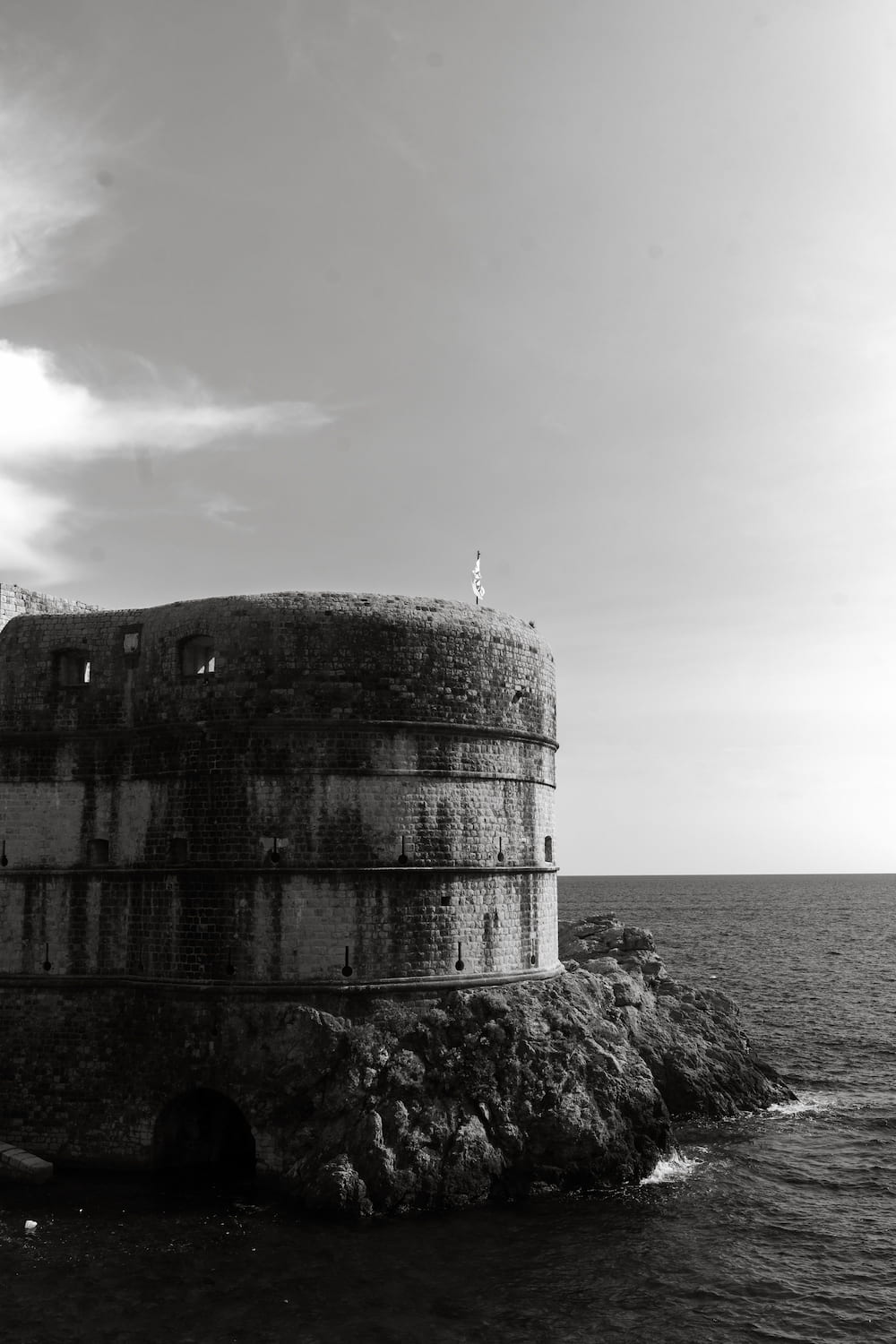 Black and white: a round stone fortress on rocks by the sea.