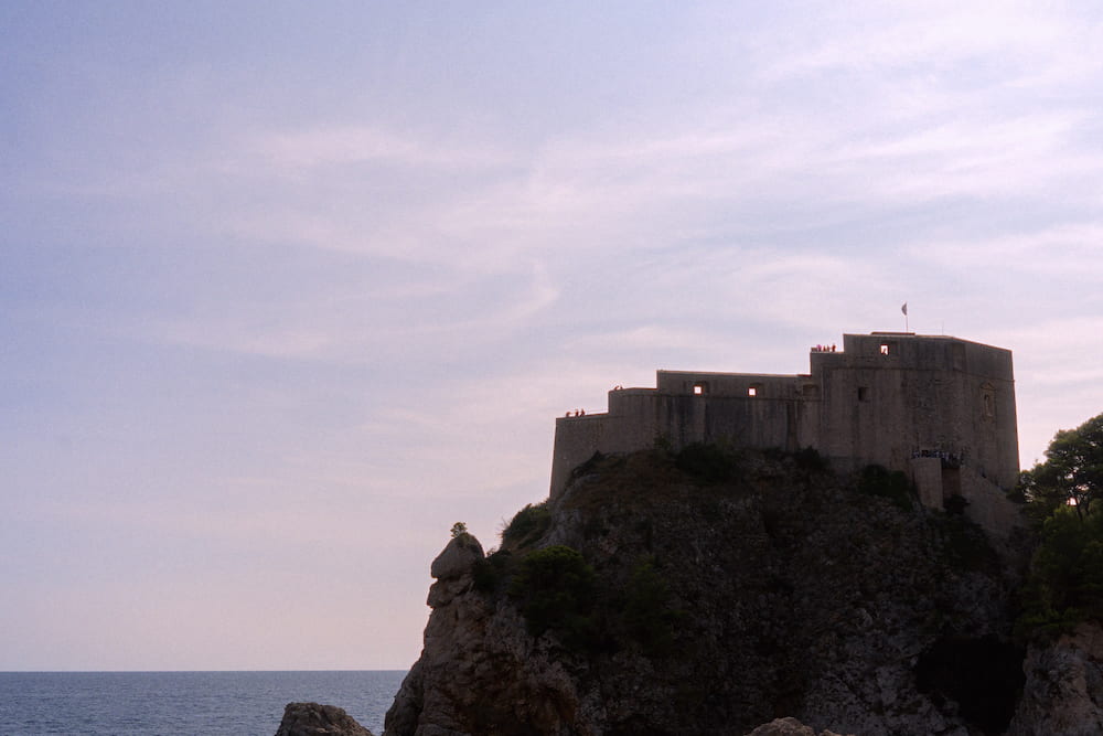 A fortress on a rock above the sea under a pink sky.
