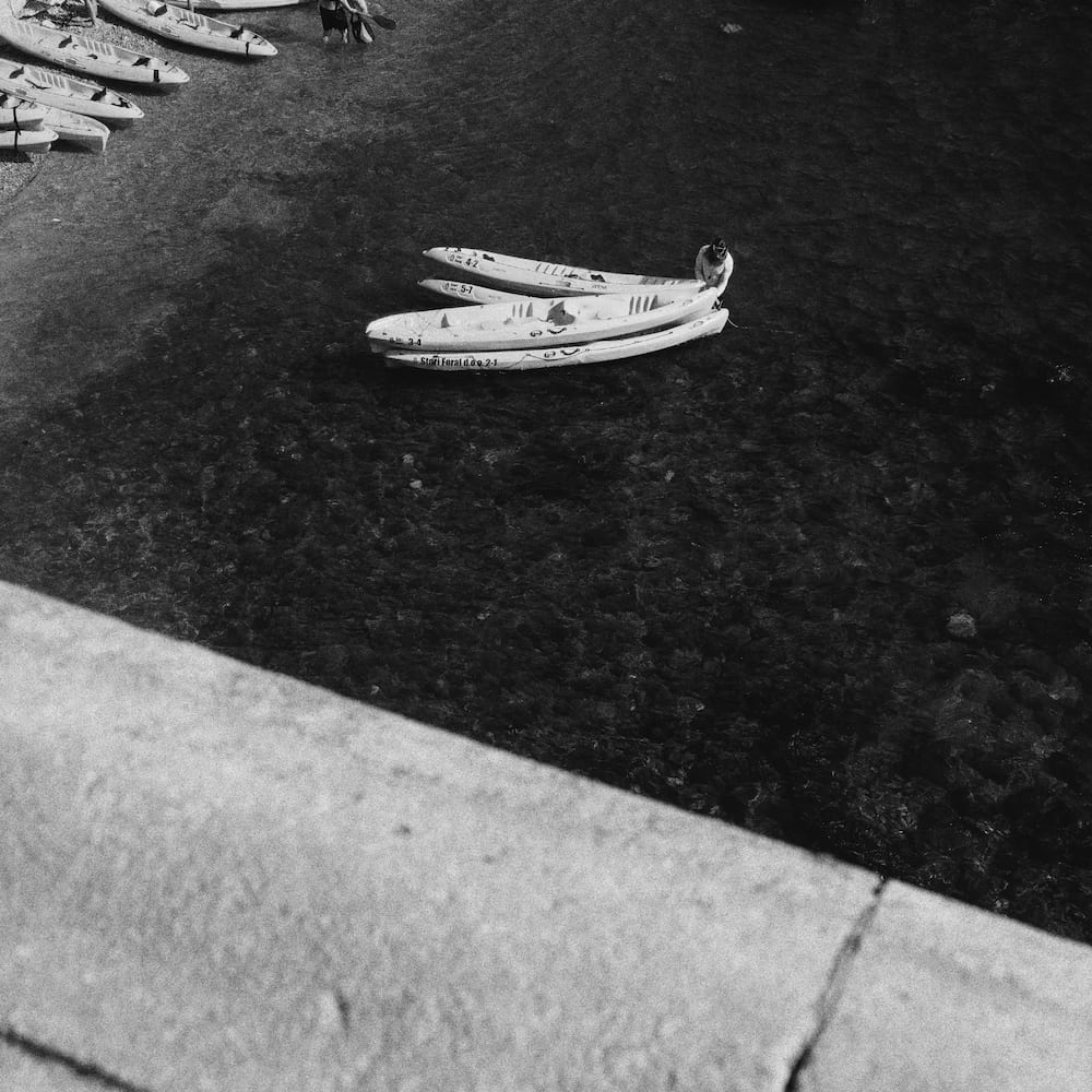 Black and white: kayaks on dark water seen from above a wall.