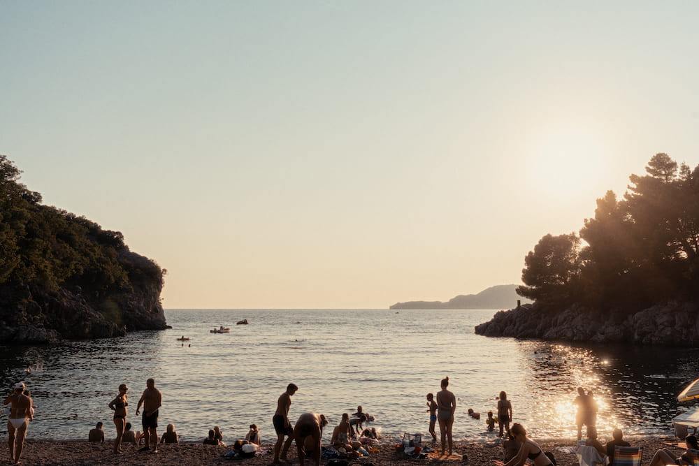 People in silhouette on a pebble beach in a cove at sunset, boats on the water.