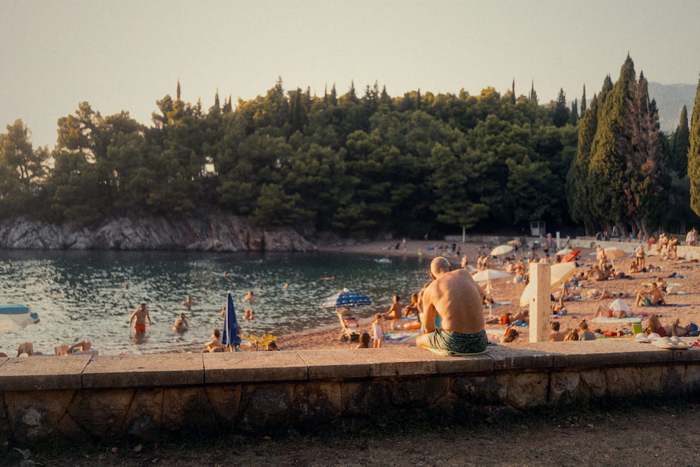 A man sitting on a wall above a crowded beach in a wooded cove.