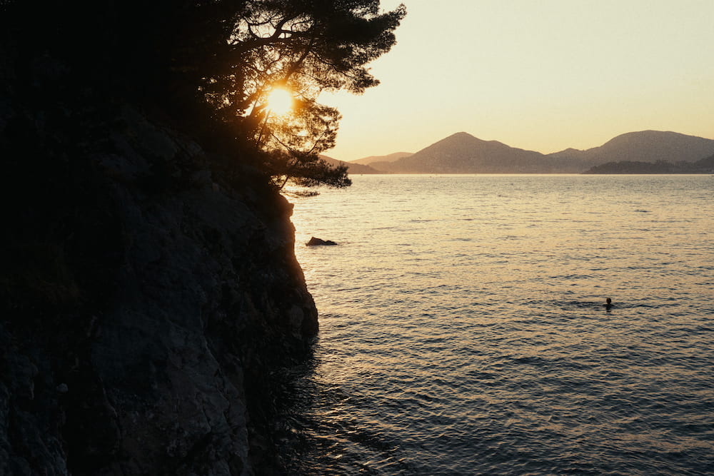 A dark cliff with trees, the sun low over the sea and hills.