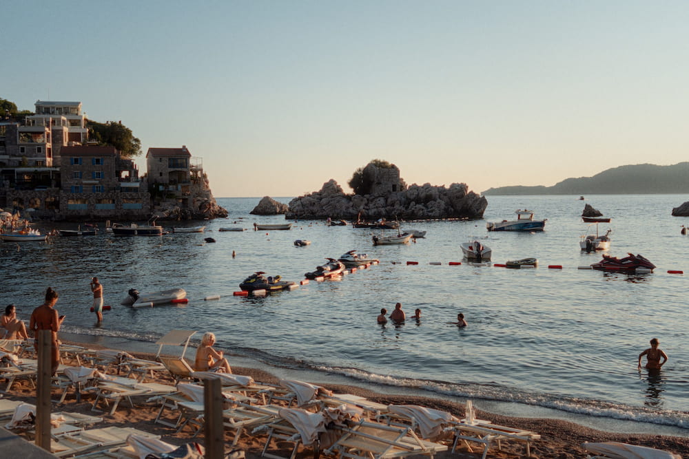 Swimmers and anchored boats in a small bay at sunset, a rocky islet behind