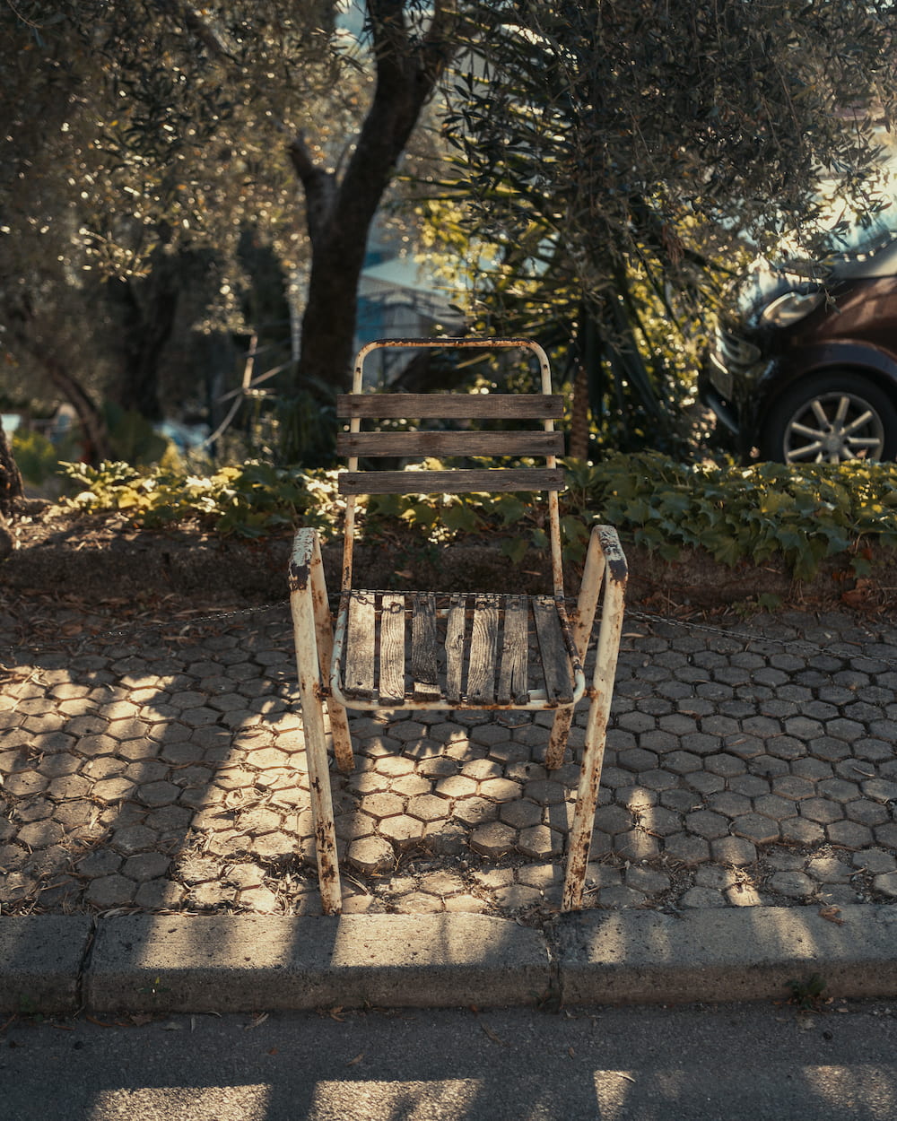 A wooden chair on paving striped with shade under olive trees