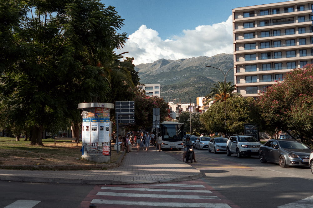 A wide street with a poster column, a hotel block and mountains behind