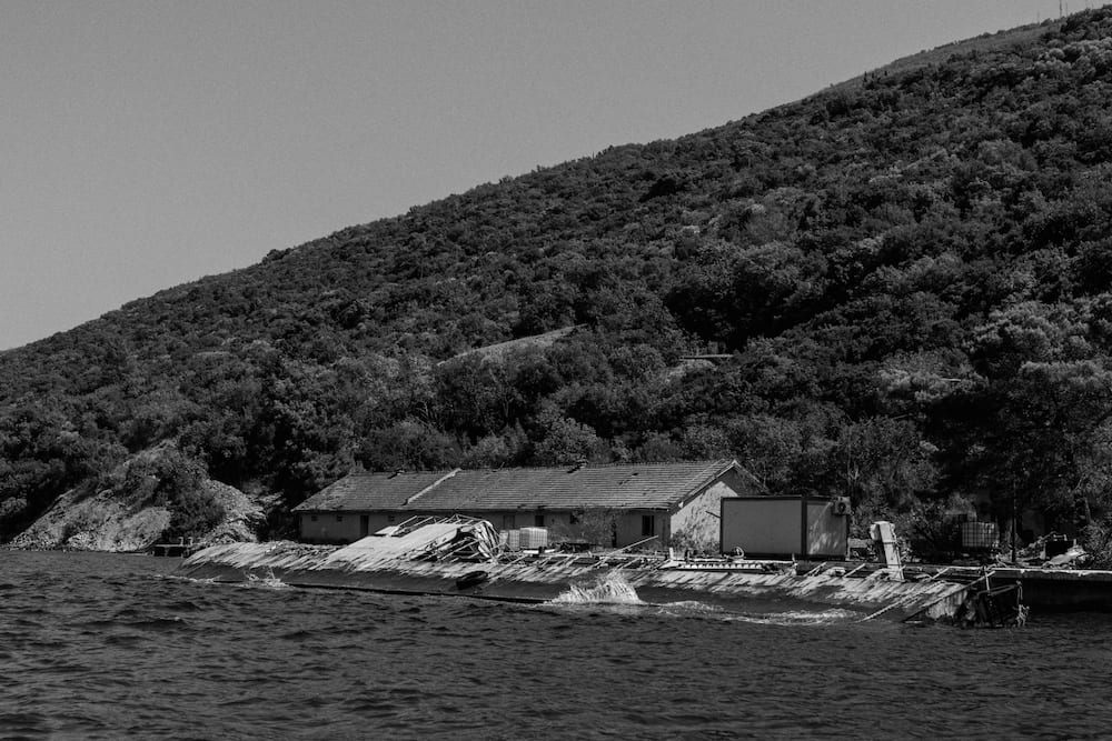 Black and white: a long low building on the shore below a wooded hill