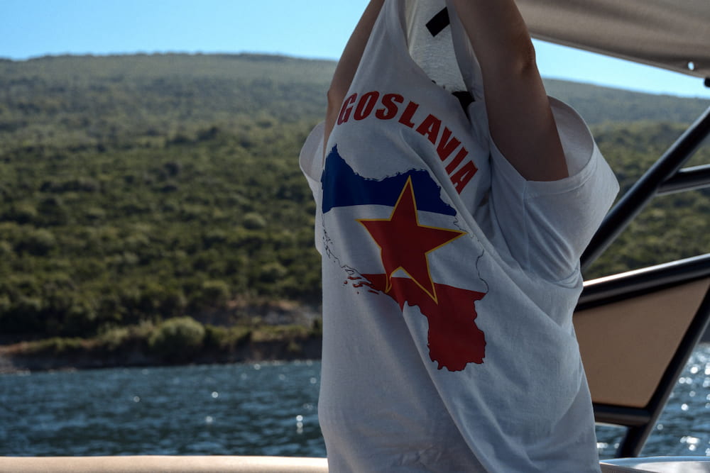 Someone in a Yugoslavia T-shirt on a boat, a green hillside behind