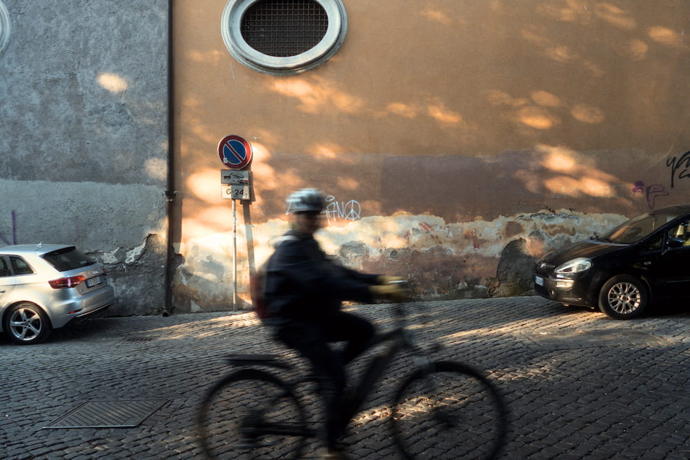 A man cycling past a pink wall with a round window, parked cars.