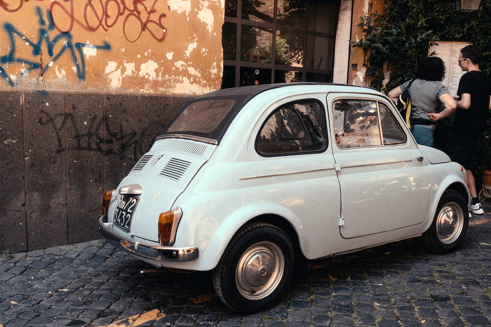 A white Fiat 500 parked on cobbles below a wall with graffiti, two people behind it.