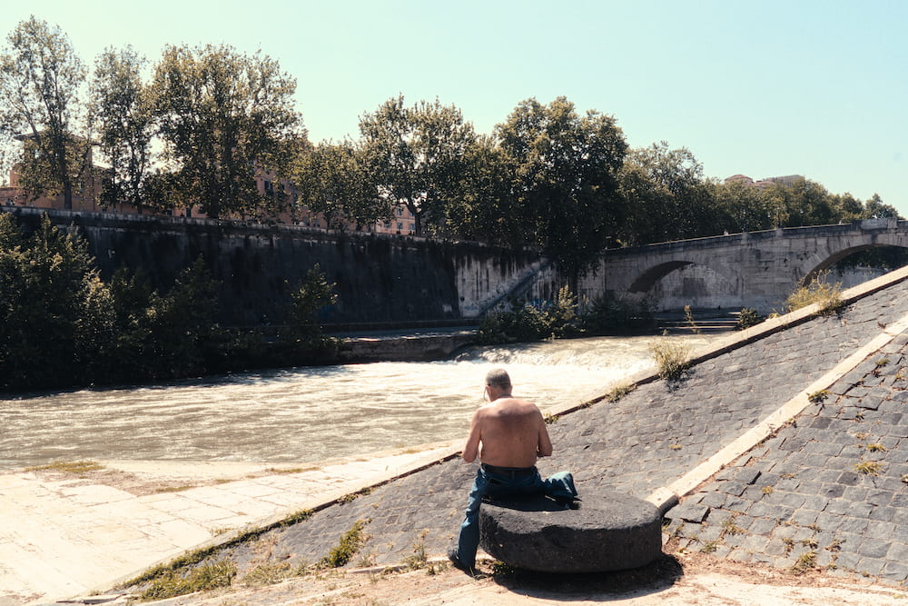 A shirtless man sitting on the stone embankment of the Tiber above a weir.