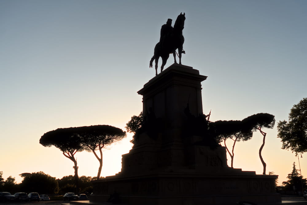 An equestrian statue on a tall plinth in silhouette, umbrella pines around it.