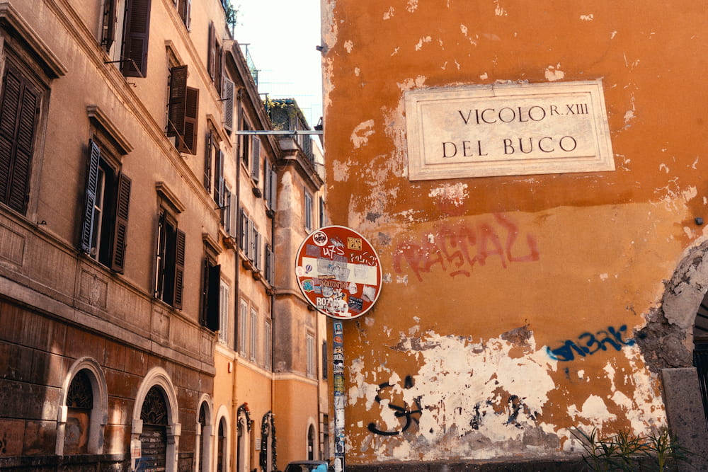 A marble street plaque, Vicolo del Buco, on a peeling orange wall beside a no-entry sign.