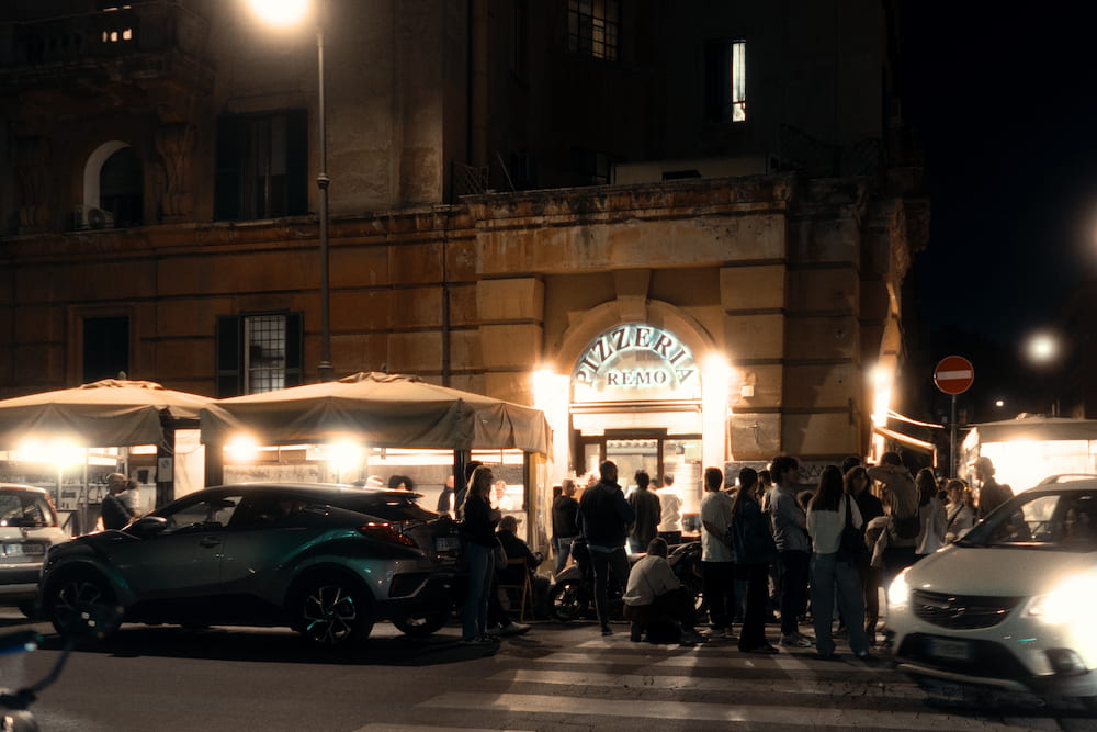 A lit restaurant terrace at night, people and parked cars in front.