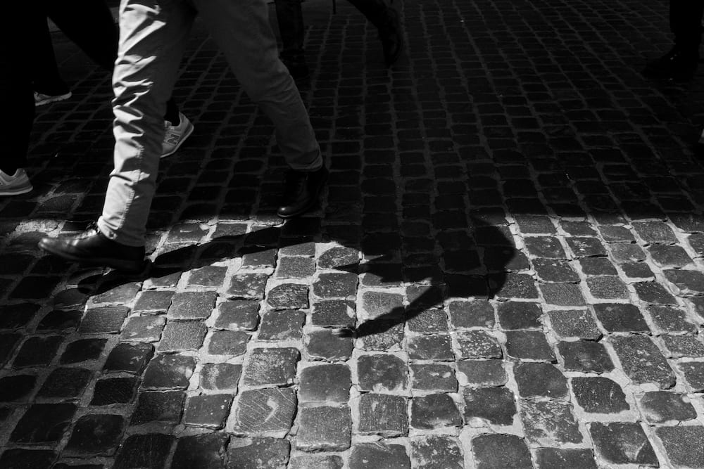 The legs of someone walking on cobbles, a hard shadow.