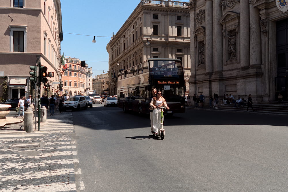 A woman on a scooter crossing a wide street, buildings around.