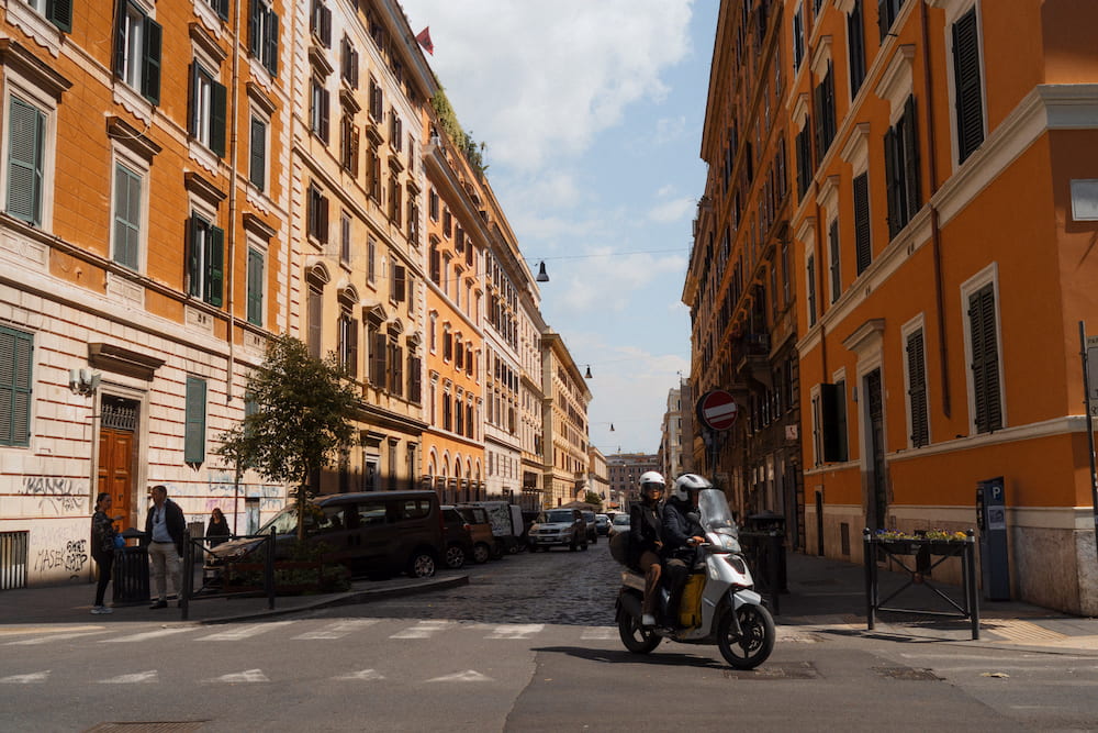 An ochre street of apartment blocks, two people on a scooter crossing.