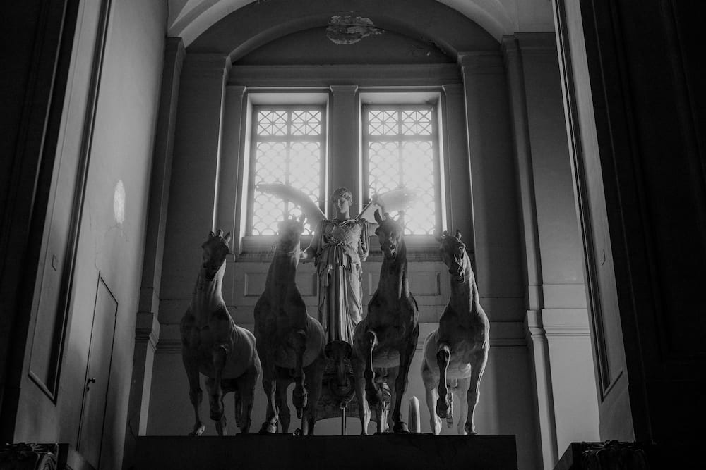 Black and white sculpture of a winged figure behind four horses, in front of tall windows.
