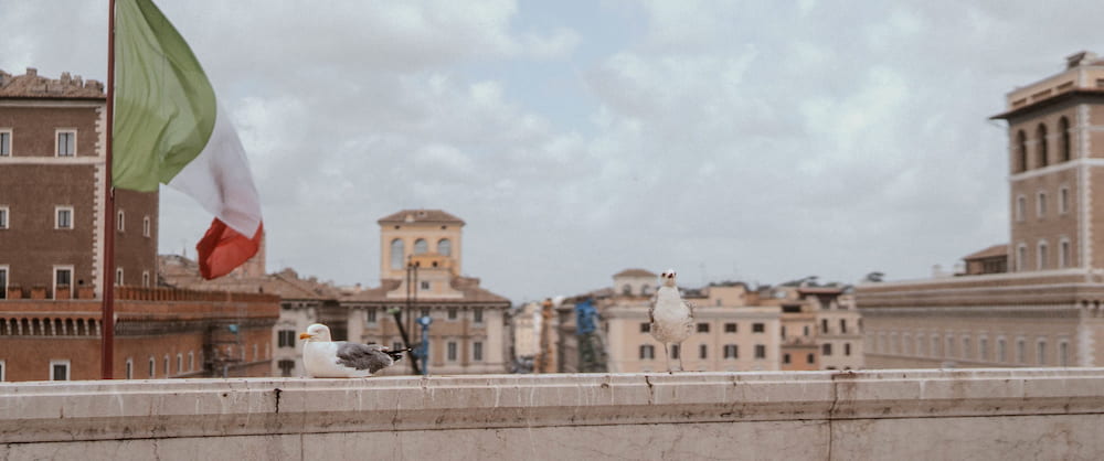 An Italian flag over the Tiber embankment and the buildings beyond, wide.