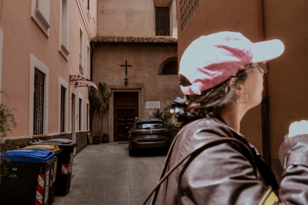 A woman in a pink cap seen from behind in a street with bins, a church front beyond.