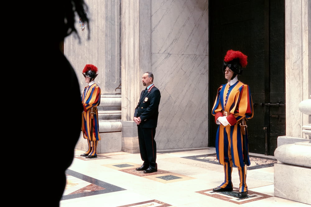 Two Swiss Guards in striped uniform and a man in a suit at a doorway, a dark figure in front.