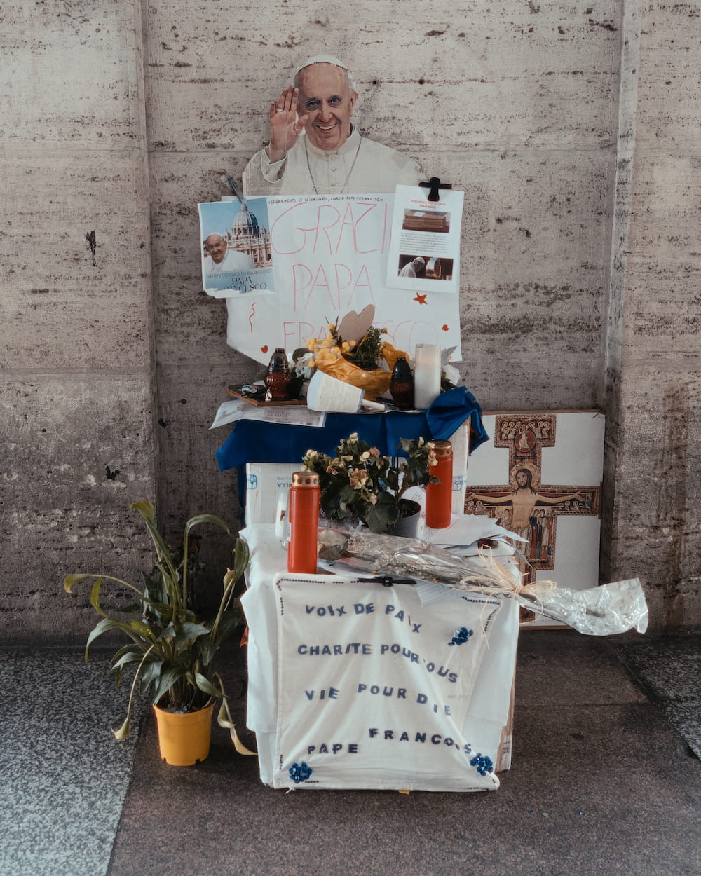A small stall against a stone wall with a picture of the Pope, flowers and drinks.