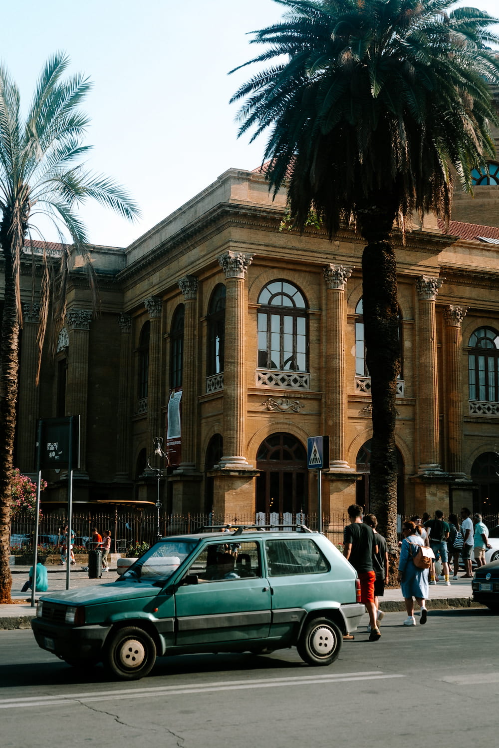 Old green hatchback passing a columned stone building, with palm trees and people on the pavement