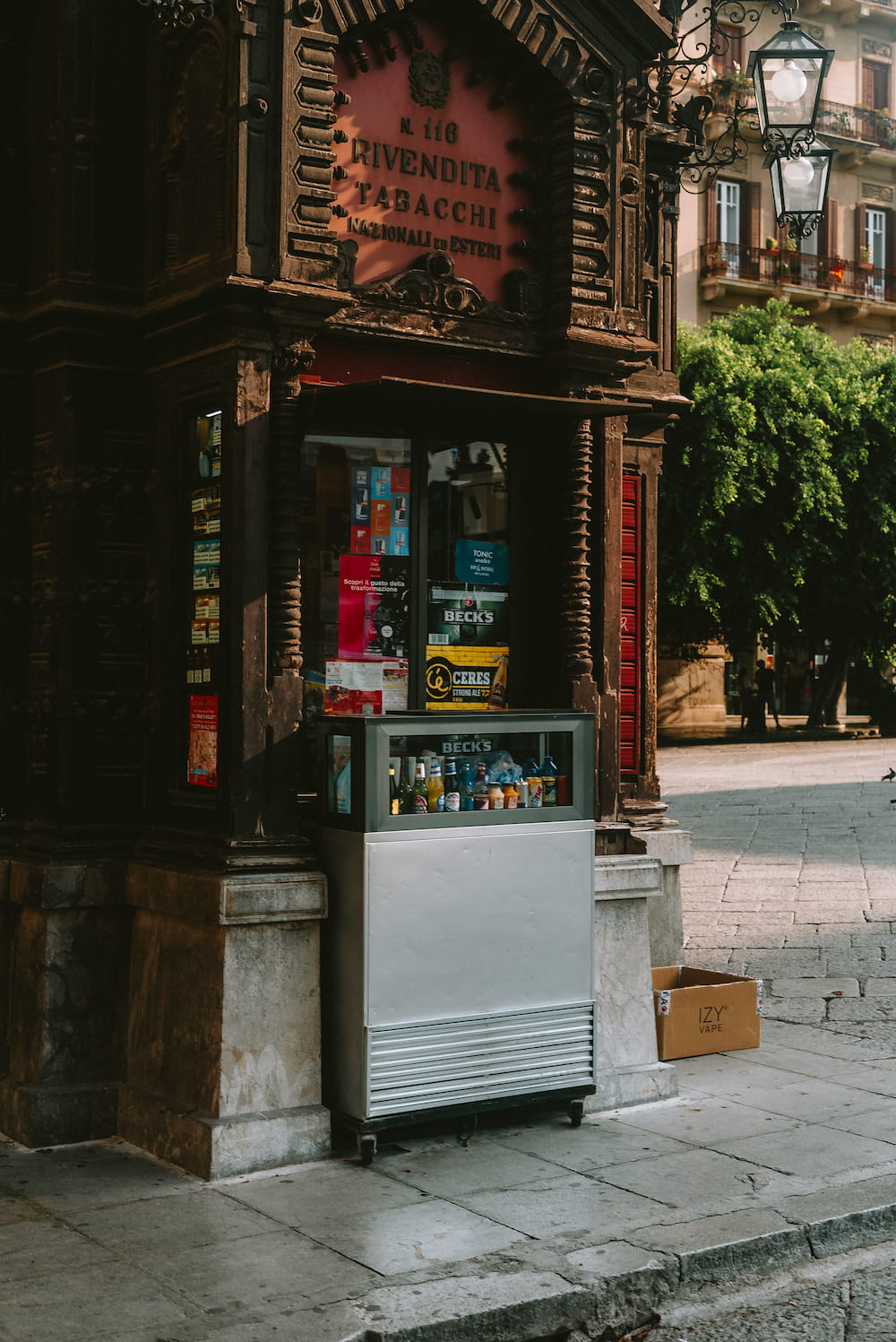 Ornate dark wooden tobacco kiosk with a red sign and a drinks fridge on the pavement