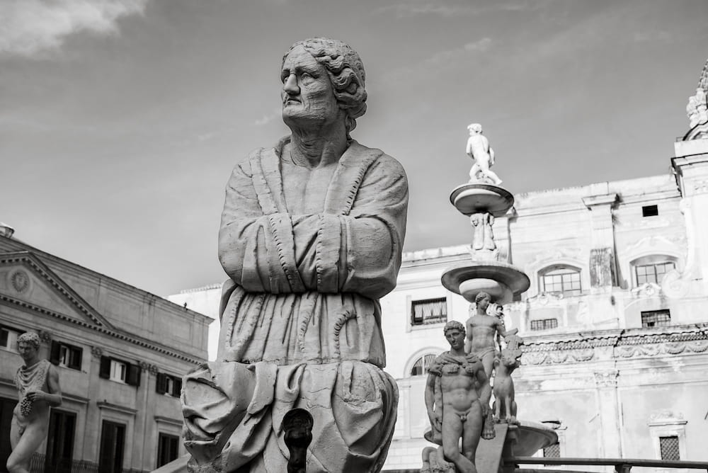 Black and white marble statue of an old man with folded arms before fountain figures and a palazzo