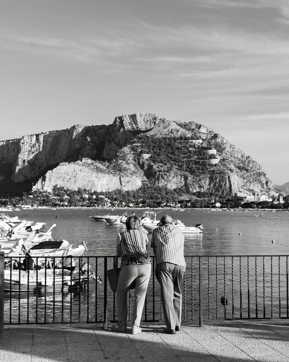 Black and white older couple leaning on a railing over a harbour of boats below a rocky mountain