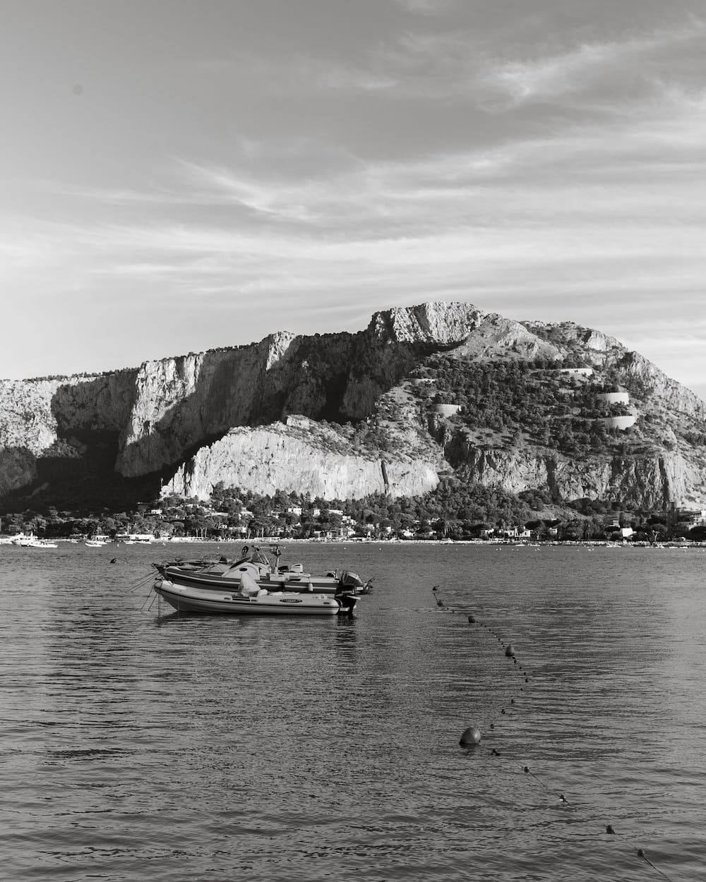 Black and white motorboats moored in a calm bay with a line of buoys below a cliff-faced mountain