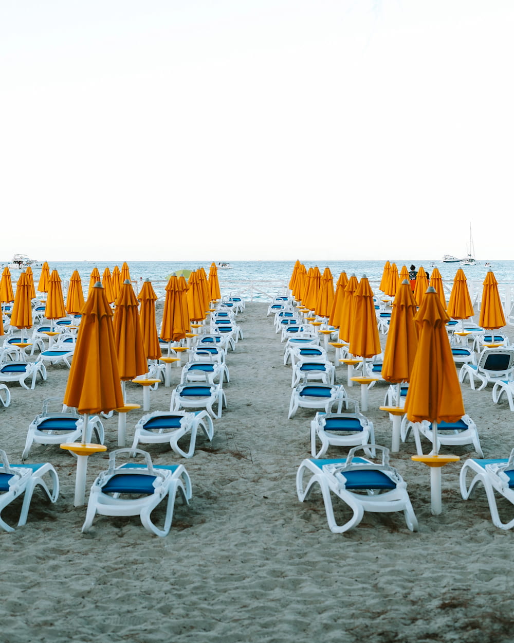 Rows of white sun loungers with blue seats and closed orange parasols on sand facing the sea