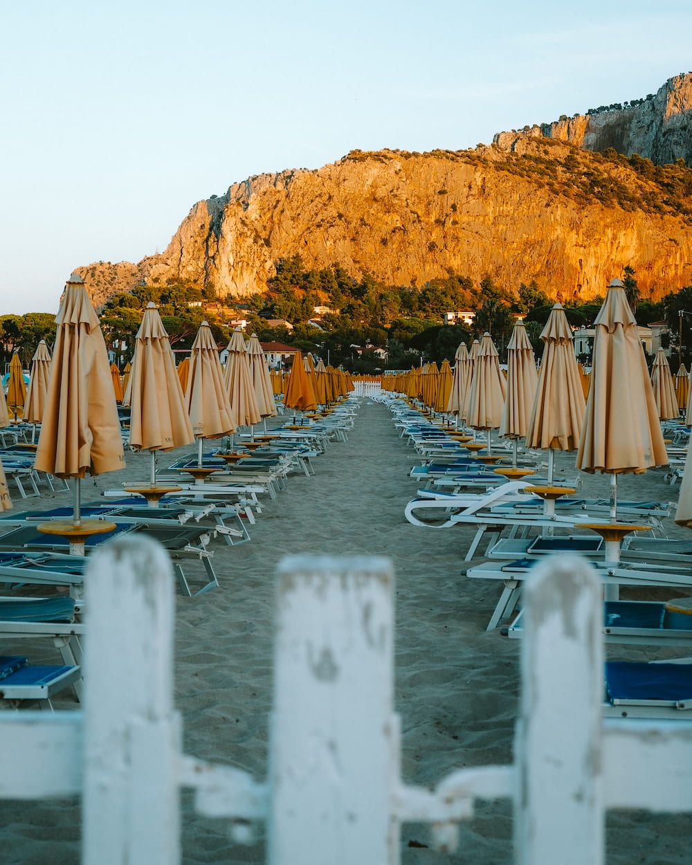 Closed beige parasols and loungers in rows behind a white fence, below a cliff lit by low sun
