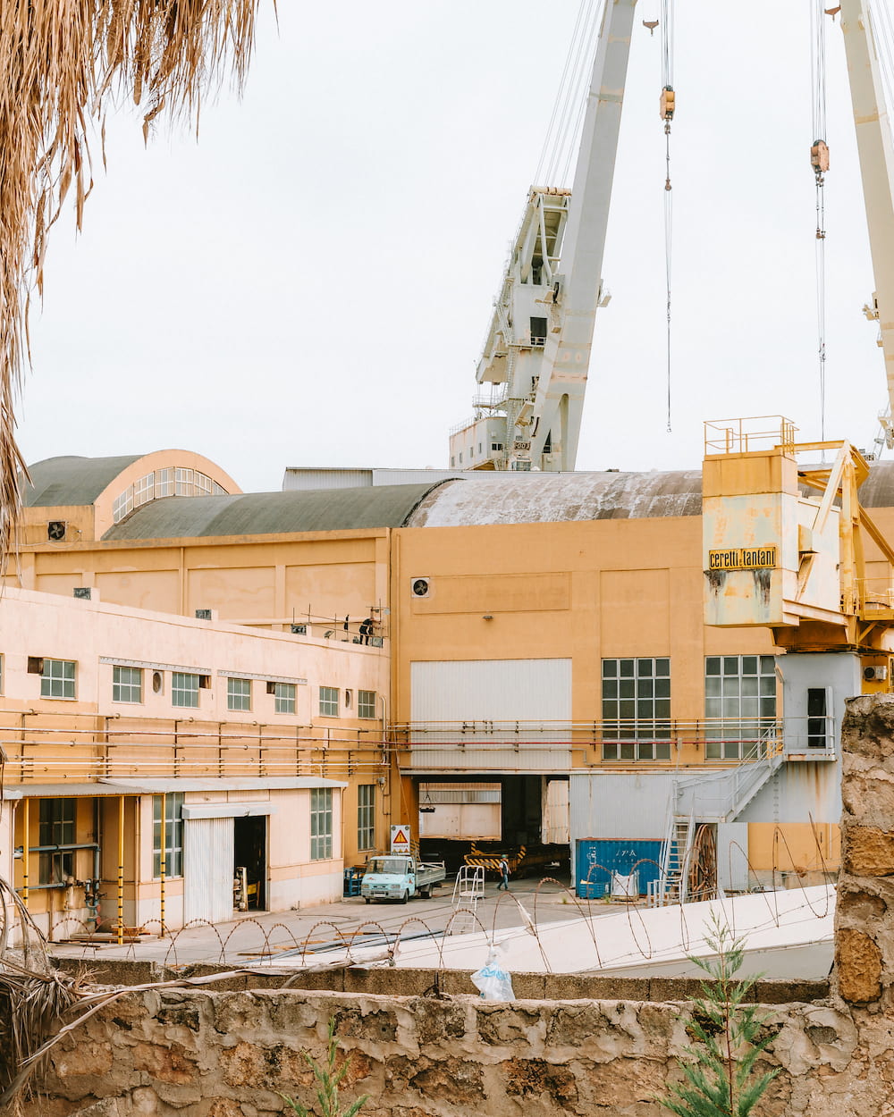 Pale orange shipyard buildings with tall cranes above, seen over a stone wall and razor wire