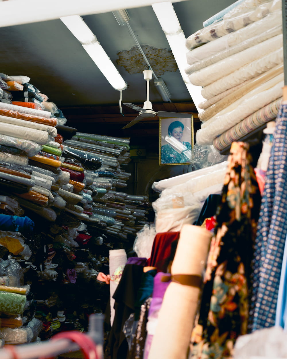 Cramped fabric shop stacked with rolls of cloth under strip lights, a framed portrait on the back wall