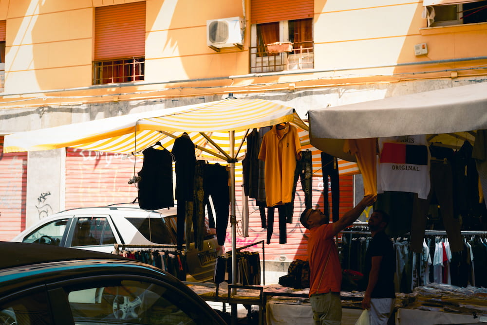 Man reaching up to clothes hanging from a striped market stall awning in front of an orange building