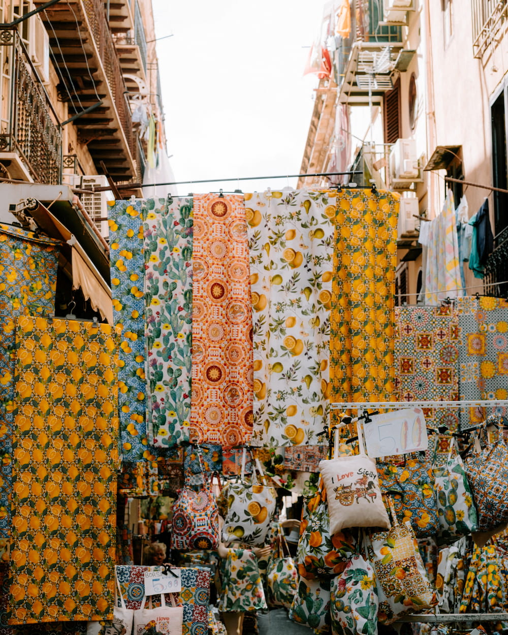 Printed tablecloths with lemons, cacti and tile patterns hung across a narrow street above tote bags
