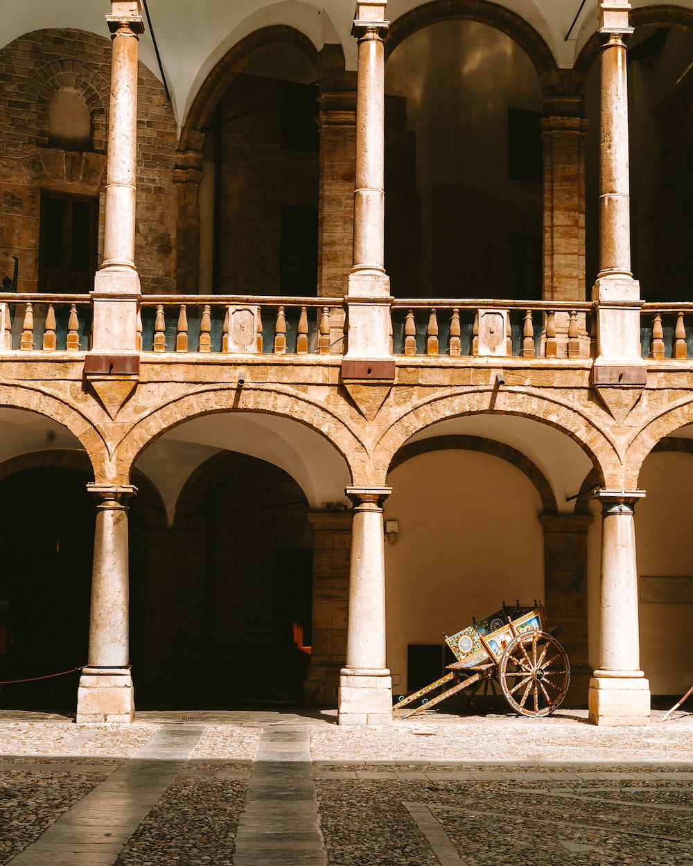 Arcaded courtyard with stone columns and a painted wooden cart standing in sunlight
