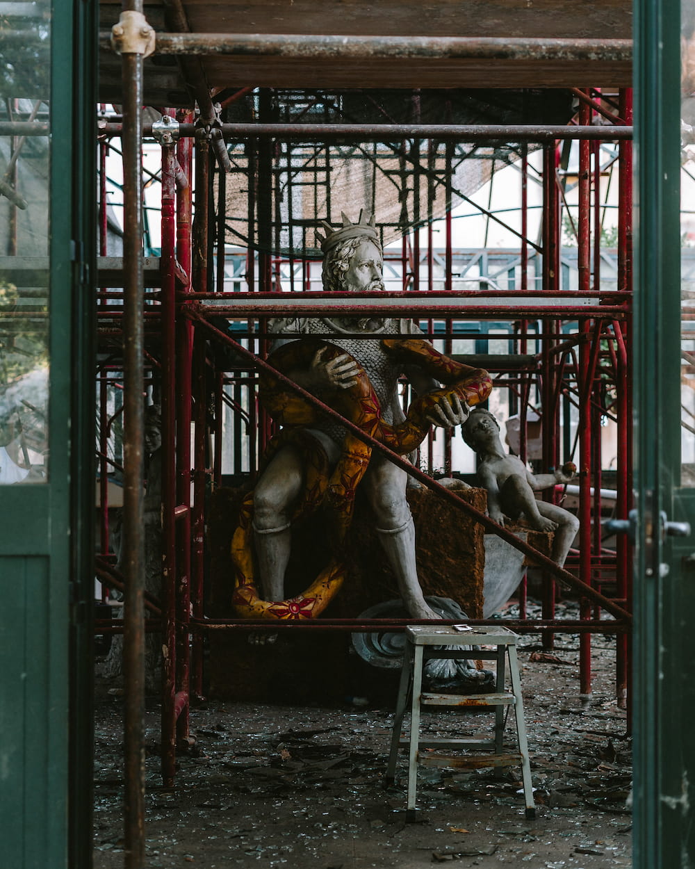 Crowned bearded statue wrapped by a painted serpent beside a cherub, inside red scaffolding with a stepladder