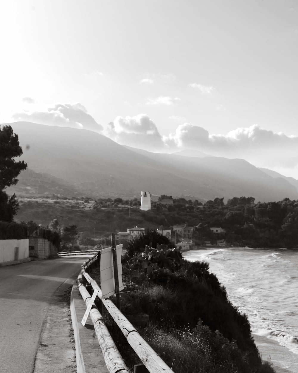 Black and white coast road with a wooden rail above a beach, a white tower and hazy hills