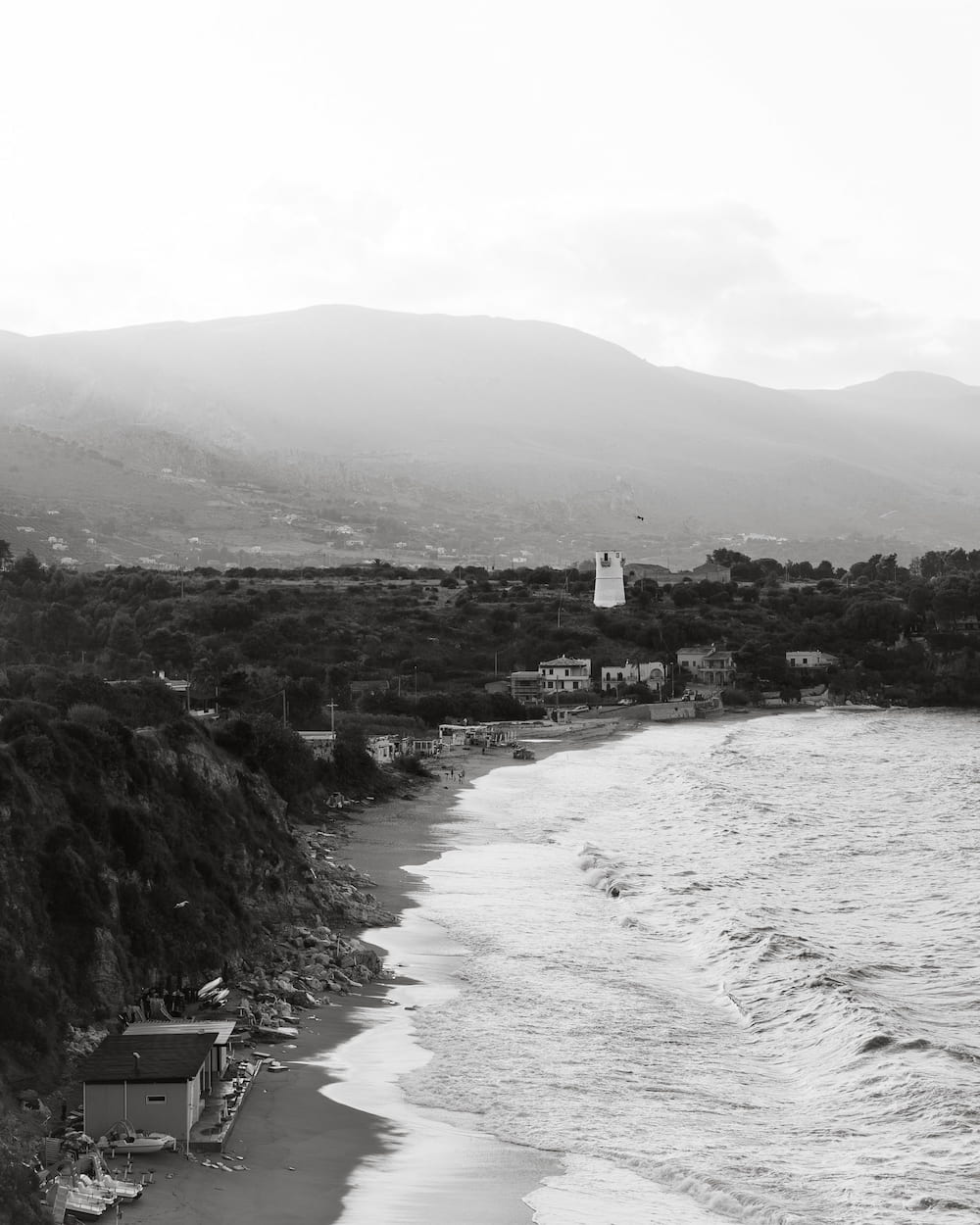 Black and white long beach with breaking waves, small huts and a white tower below misty hills
