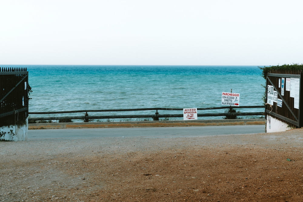 Open gate onto a road and wooden fence with beach signs, a turquoise sea beyond