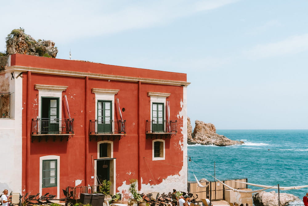Red house with green shutters and iron balconies beside a rocky outcrop and turquoise sea