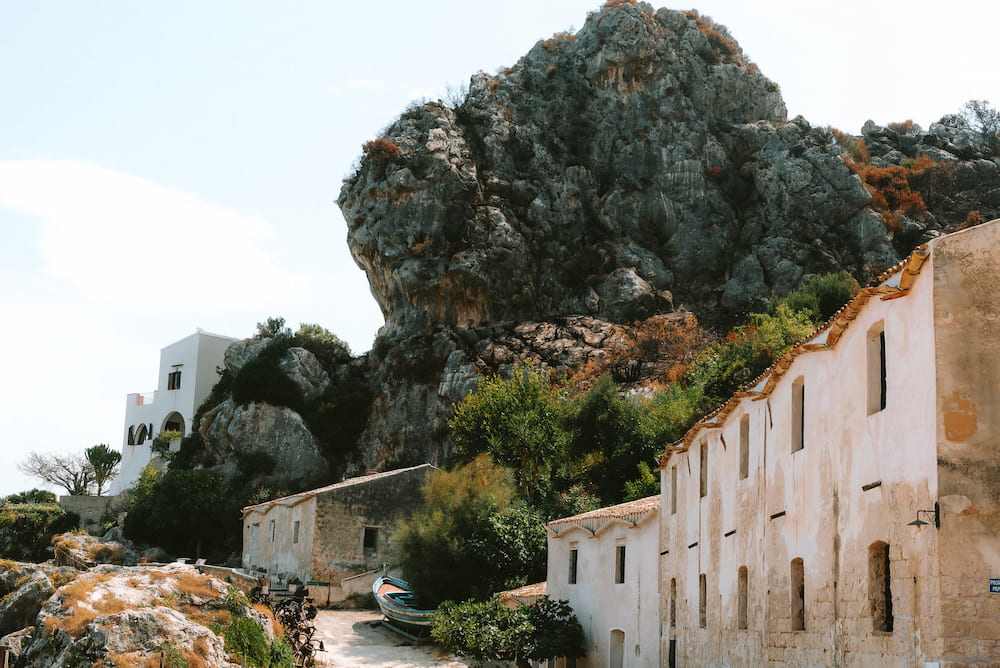 Old whitewashed buildings and a small boat on a sandy lane below a huge grey rock