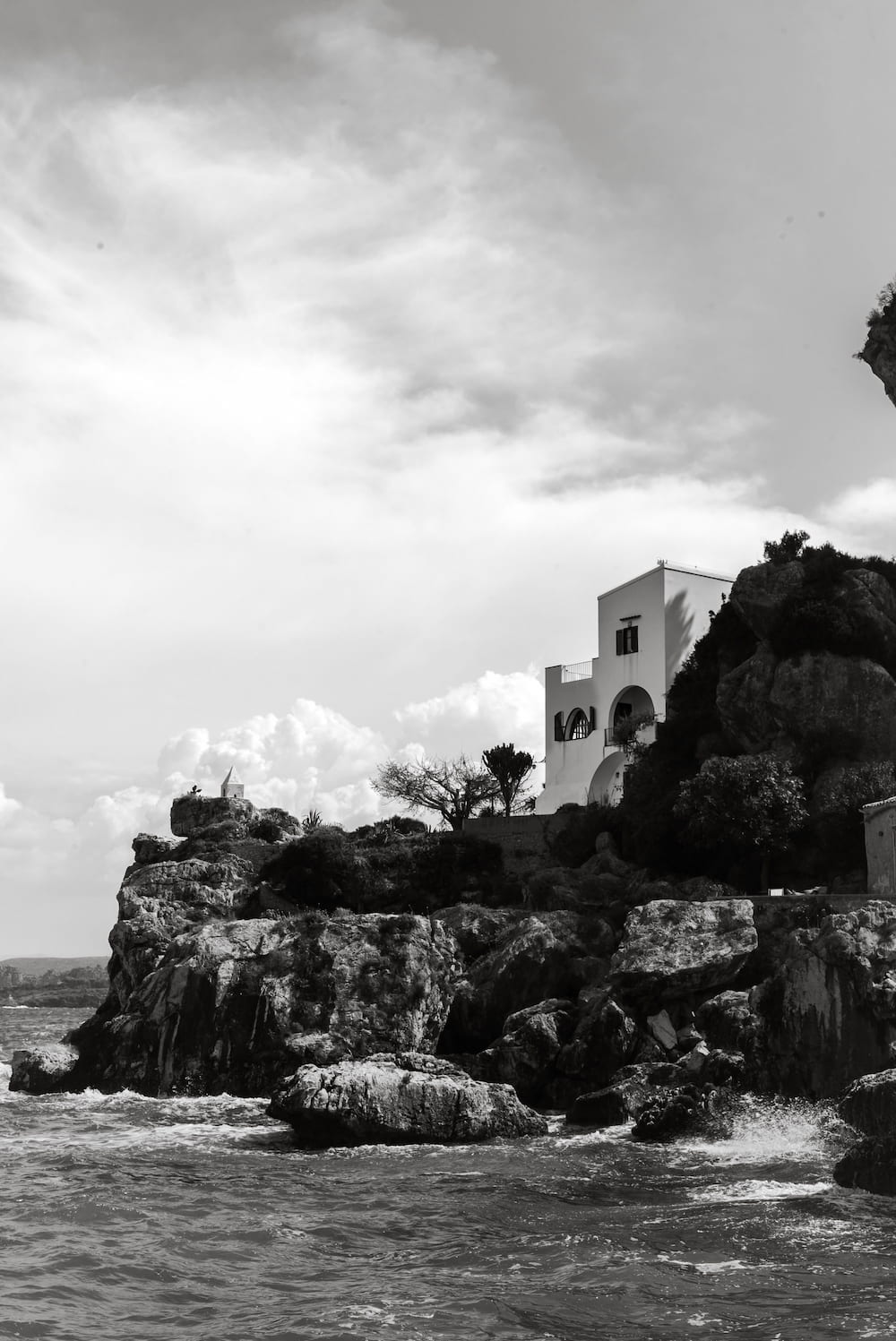 Black and white villa on rocks above a choppy sea under a bright cloudy sky