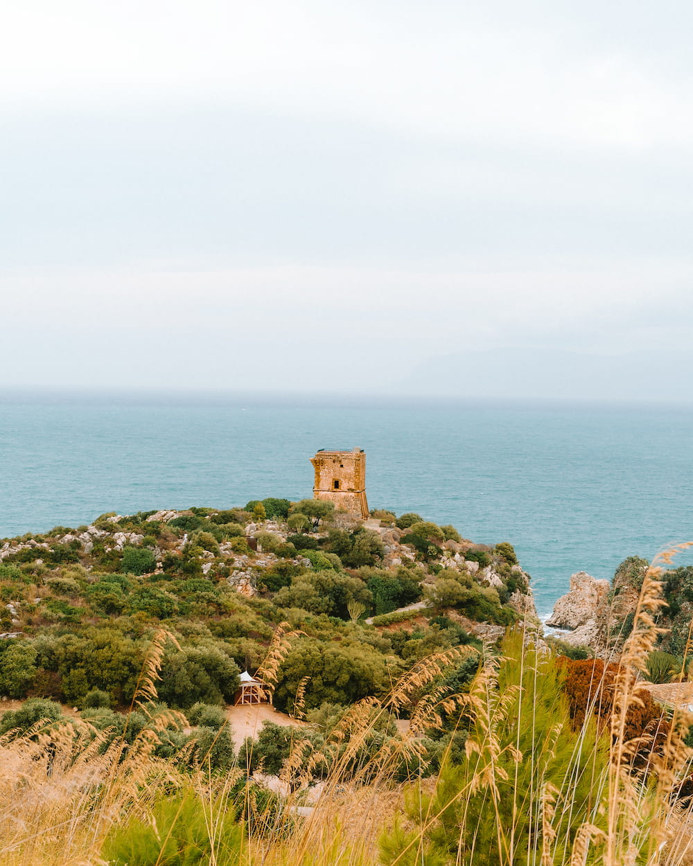 Square stone tower on a scrubby headland above the sea, seen past tall dry grass