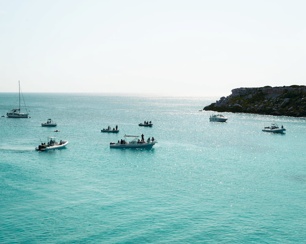 Small motorboats and a sailboat anchored in turquoise water near a low rocky point