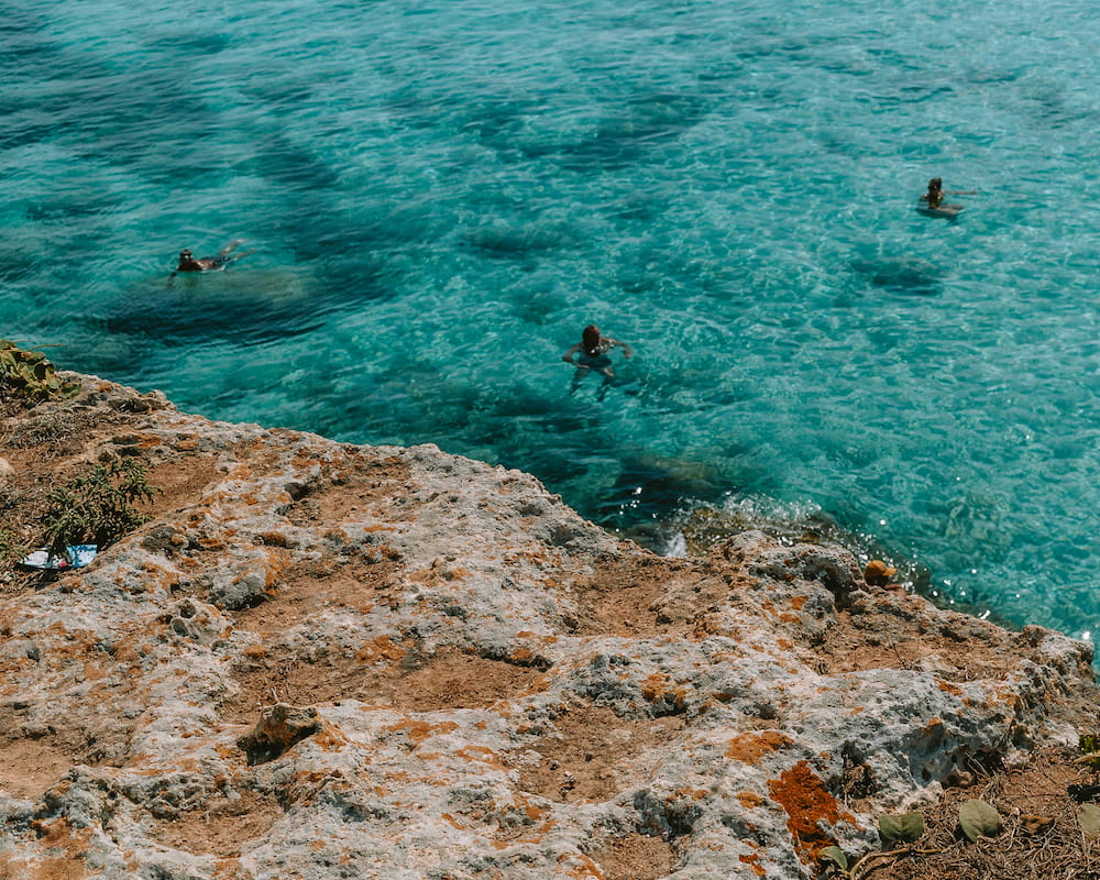 Three swimmers in clear turquoise water seen from a lichen-covered rock ledge above