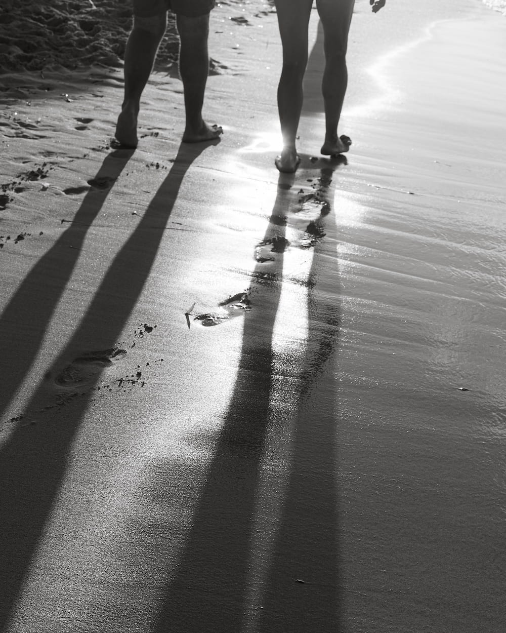 Black and white two people walking barefoot on wet sand, their long shadows stretching back