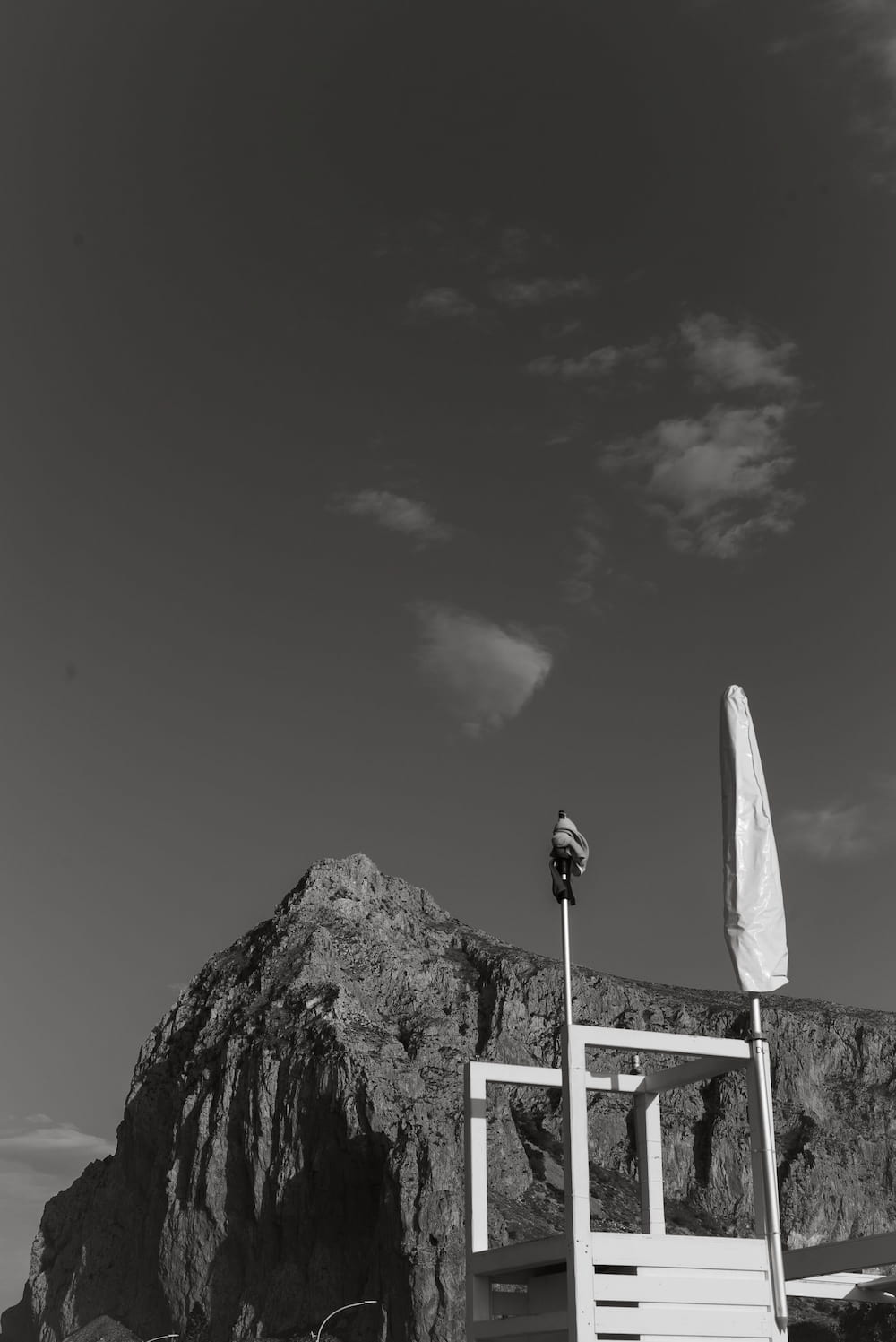 Black and white lifeguard tower with a furled umbrella before a steep rock peak and dark sky